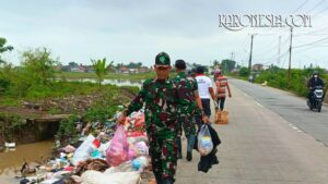 Personel TNI Koramil 08/Kronjo mengenakan seragam loreng sedang mengangkat kantong plastik berisi sampah di pinggir jalan Desa Muncung.