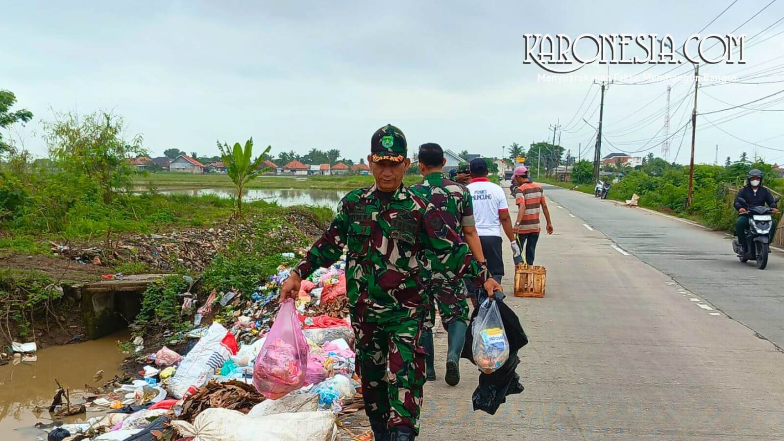 Personel TNI Koramil 08/Kronjo mengenakan seragam loreng sedang mengangkat kantong plastik berisi sampah di pinggir jalan Desa Muncung.