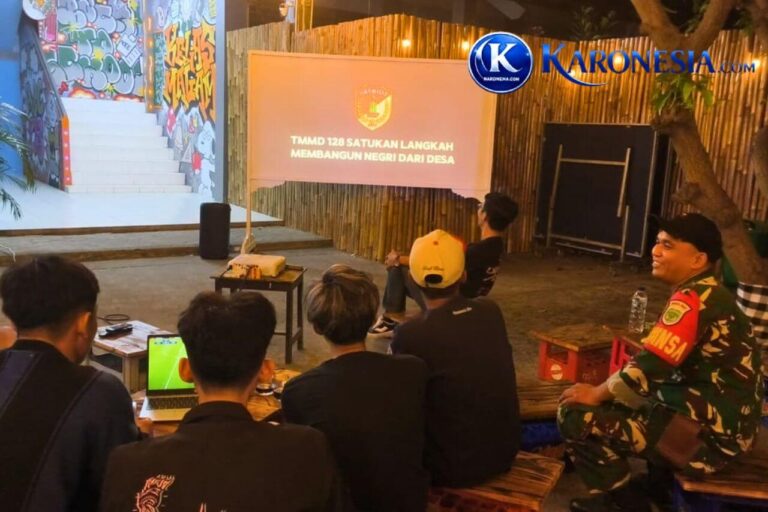 Babinsa berpakaian dinas duduk bersama para pemuda menyaksikan tayangan di layar proyektor.