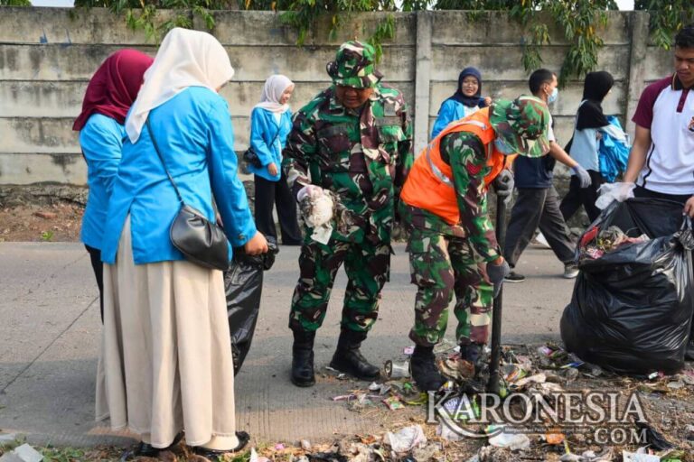 Personel TNI berbaju loreng bersama mahasiswa berjaket biru dan masyarakat sedang memunguti sampah dan memasukkannya ke kantong plastik hitam besar. Salah satu anggota TNI menggunakan sekop untuk mengangkat tumpukan sampah di pinggir jalan. Kegiatan ini berlangsung di kawasan Desa Pasirnangka, Tigaraksa, Kabupaten Tangerang.