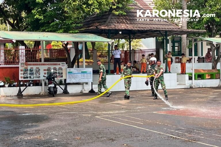 Empat prajurit TNI berseragam loreng menyemprotkan air menggunakan selang panjang di halaman sekolah, dengan siswa berseragam oranye terlihat di latar belakang.